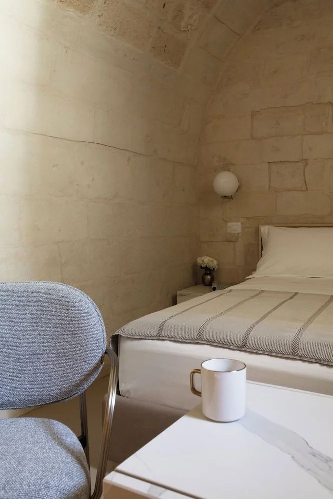 Helles Hotelzimmer mit Steinwand, weißem Doppelbett und Stuhl im Vordergrund.