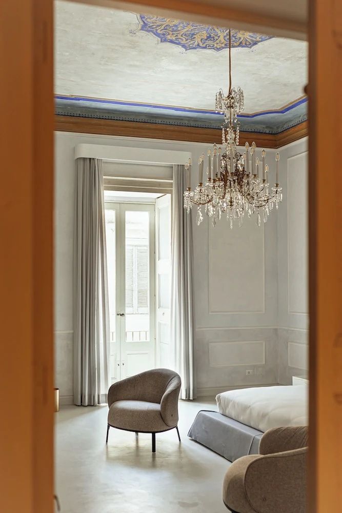 Blick in ein helles Zimmer mit bodentiefem Fenster, Vorhängen, Sessel und großem Kronleuchter.