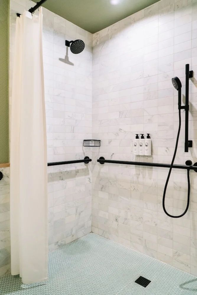Badezimmer mit gefliester Dusche, schwarzen Armaturen und Haltegriffen an den Wänden.
