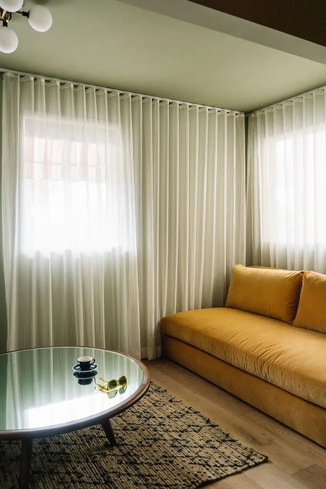 Sitzecke im Hotelzimmer mit gelbem Sofaelement, weichen Vorhängen und rundem Tisch.