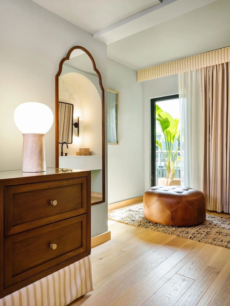 Zimmerecke mit Holzkommode, wandmontiertem Spiegel, Leder Pouf und Fensterfront.
