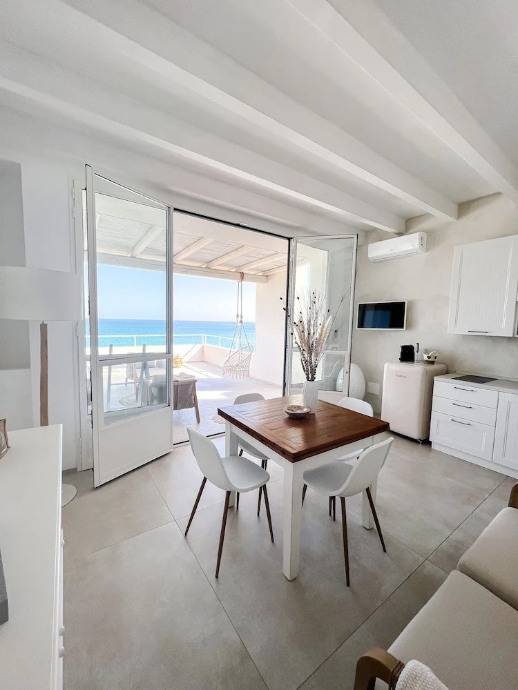 Heller Wohnbereich mit Holztisch, Küchenzeile und Blick durch die Balkontür auf das Meer.