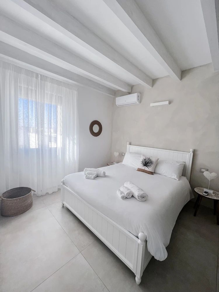 Schlafzimmer mit weißem Doppelbett, Deckenbalken, Nachttischen und vorhangverhängtem Fenster.