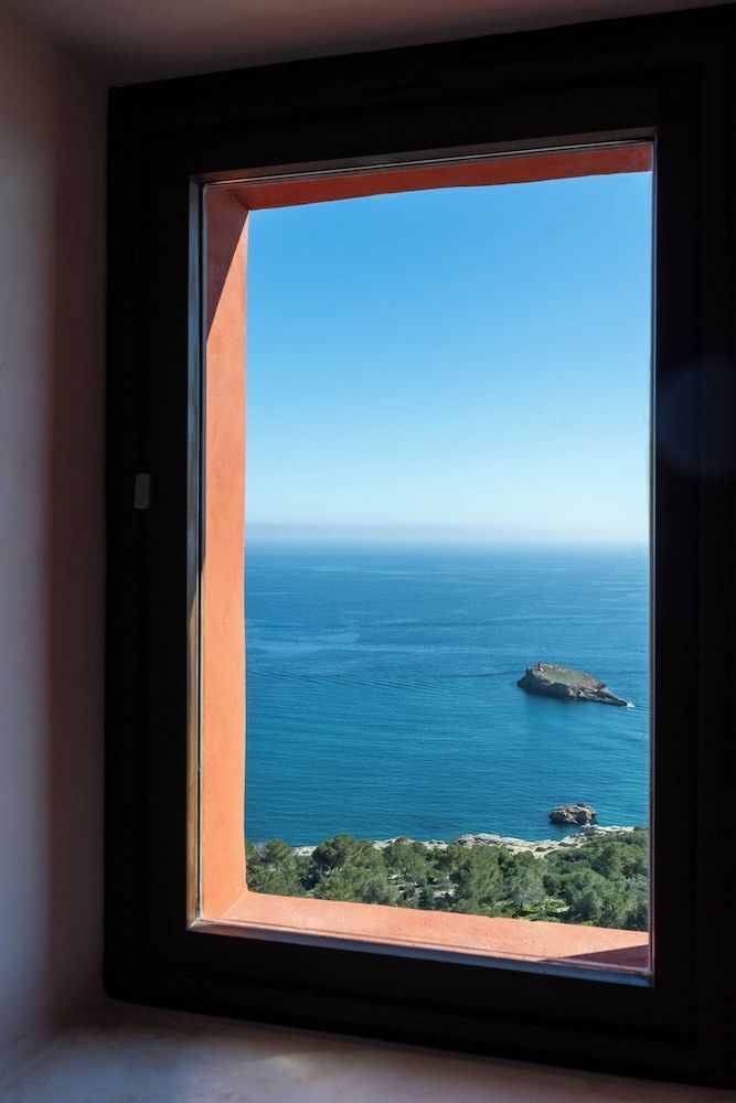 Blick durch einen lachsfarbenen Fensterrahmen auf das blaue Meer und eine kleine Insel vor der Küste.