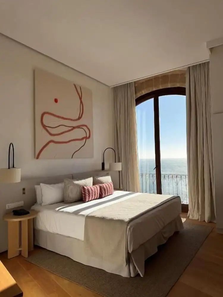 Heller Hotelraum mit einem großen Doppelbett, abstrakter Wandkunst und einem Rundbogenfenster mit Meerblick.