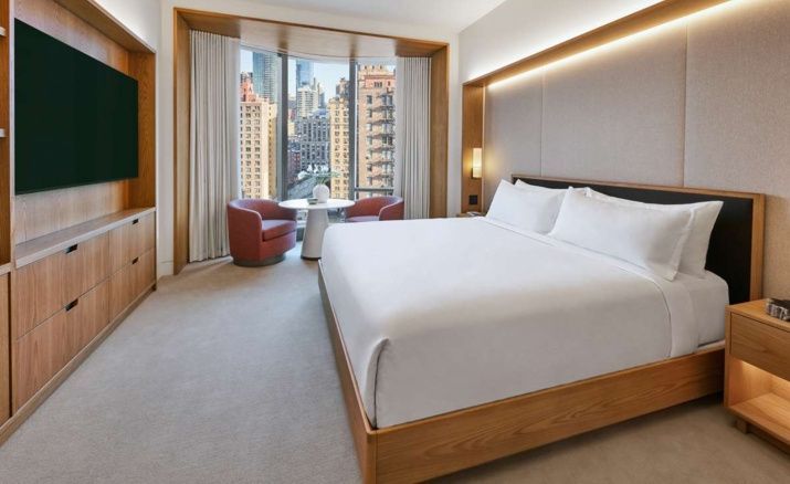 Modernes Hotelzimmer mit Holzdoppelbett, zwei roten Sesseln am Fenster und Blick auf Stadtgebäude.