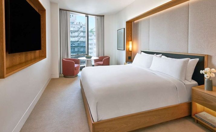Helles Schlafzimmer mit einem Doppelbett, Sitzbereich am Fenster und Blick auf Stadtgebäude.