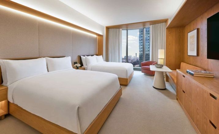 Hotelzimmer mit zwei Doppelbetten, Sessel, Tisch und Fenster mit Ausblick auf die Stadt.