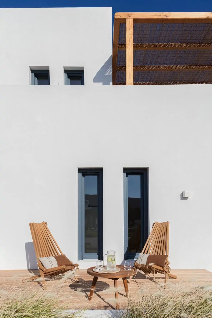 Weiße Fassade mit zwei dunklen Fenstern und Holzstühlen auf der Terrasse.