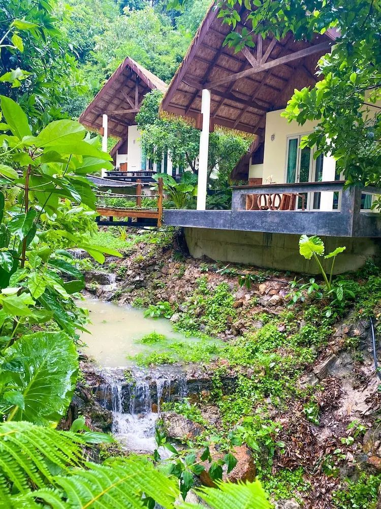 Bungalows mit Reetdächern an einem kleinen Bachlauf umgeben von grüner Tropenvegetation.
