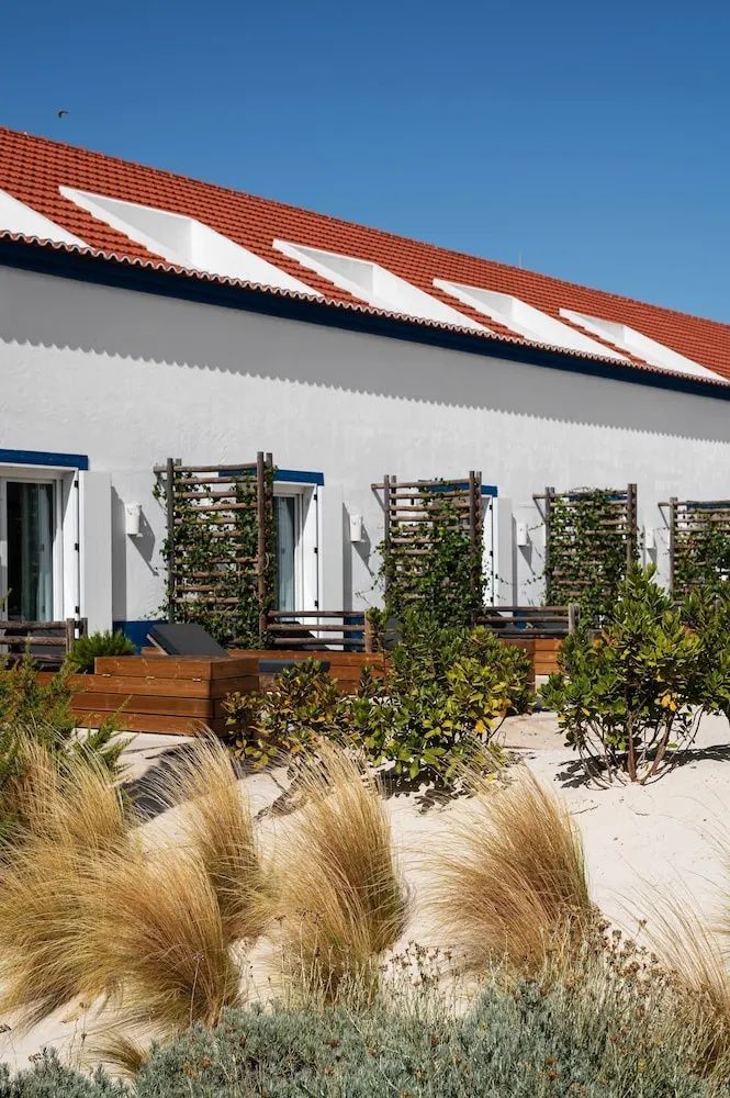 Weißes Gebäude mit rotem Ziegeldach, kleinen Holzterrassen und Bepflanzung auf Sand.