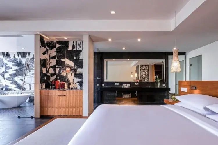 Hotelzimmer mit weißem Bett im Vordergrund und dunklem Waschtischbereich dahinter.