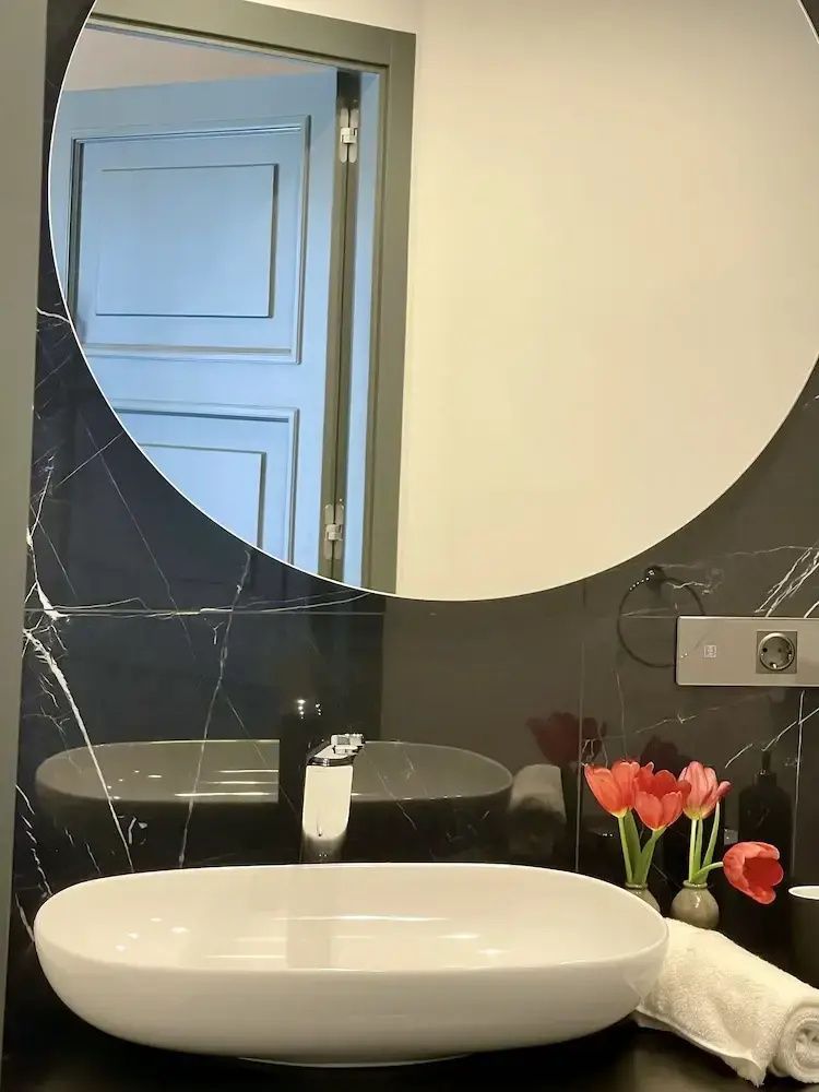 Badezimmer mit weißem Aufsatzwaschbecken, rundem Spiegel und dunklen Fliesen an der Wand.