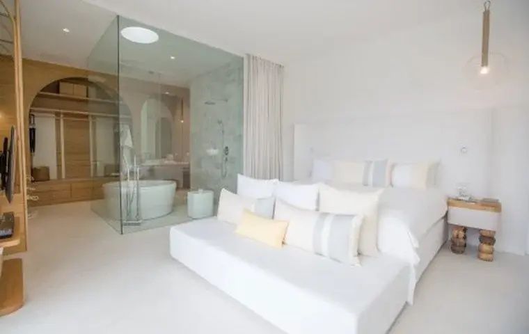 Weißes Hotelzimmer mit Doppelbett, Tagesbett und durch eine Glaswand abgetrenntem Bad mit Badewanne.