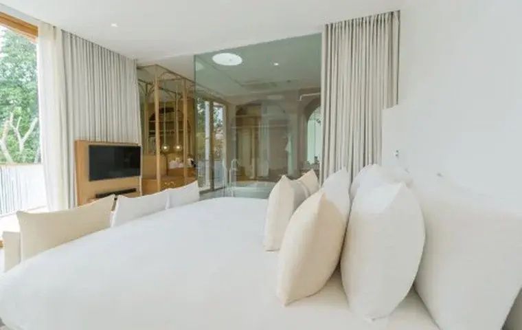 Blick über das weiße Bett hin zu Fernseher, Holzschrank und der transparenten Badabtrennung.