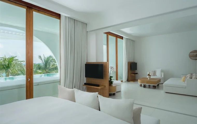 Helles Hotelzimmer mit weisem Bett, Fernseher auf Raumteiler und Sitzbereich mit Fenstern.