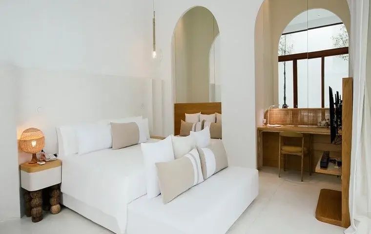 Weißes Hotelzimmer mit Doppelbett, beigen Kissen und holzverkleidetem Arbeitsbereich.