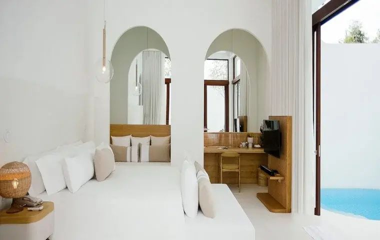 Helles Schlafzimmer mit weißem Bett, Rundbögen an der Wand und Arbeitsplatz mit Stuhl.