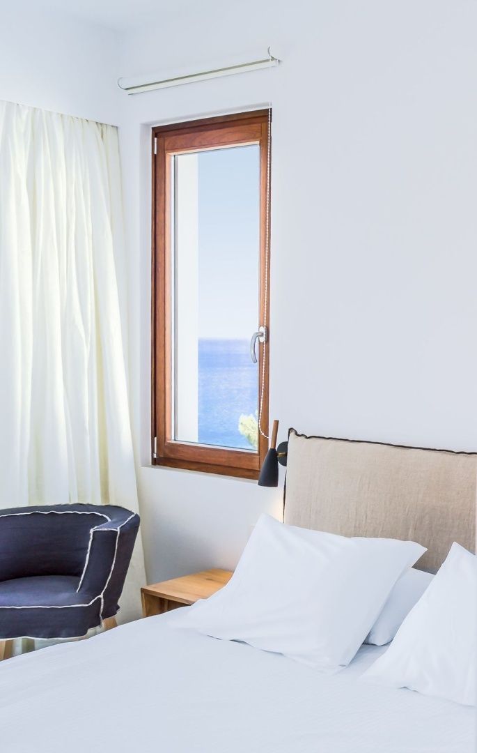 Blick aus dem Fenster eines hellen Hotelzimmers auf das blaue Meer im Sonnenschein.