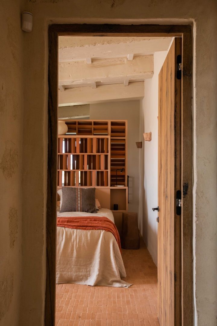 Blick durch eine Holztür auf ein Bett mit oranger Decke und Holzregal dahinter.