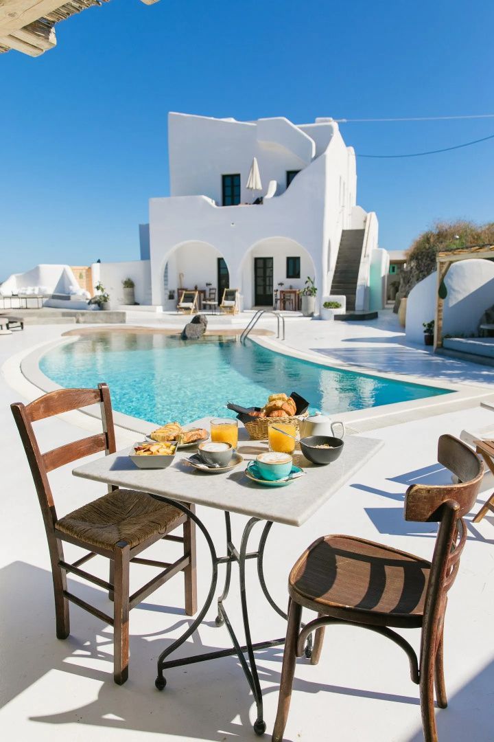 Gedeckter Frühstückstisch am Swimmingpool vor weißem Gebäude im kykladischen Stil.