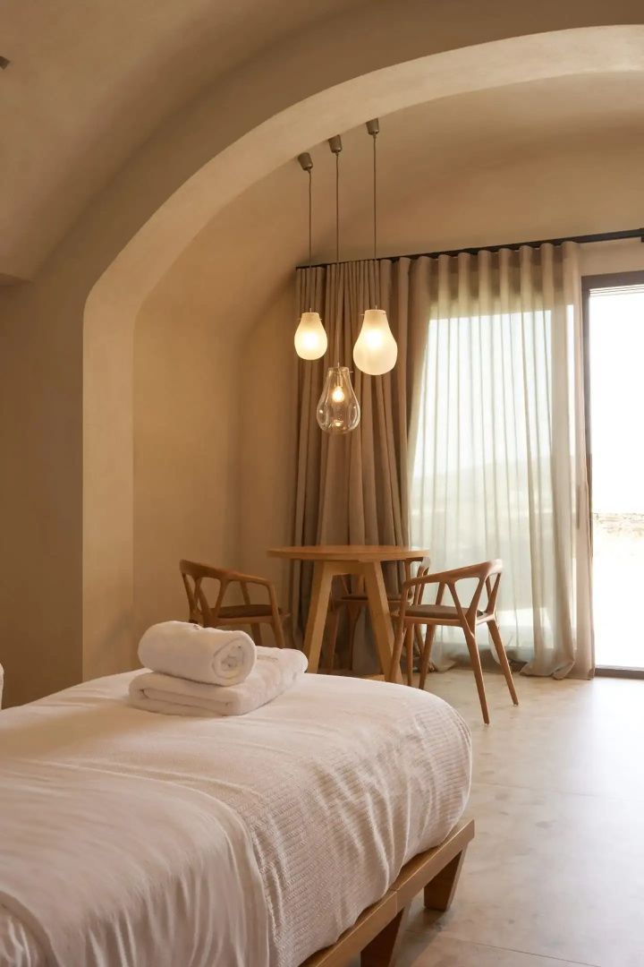 Hell eingerichtetes Schlafzimmer mit Holztisch, Stühlen und Bett mit weißen Handtüchern.