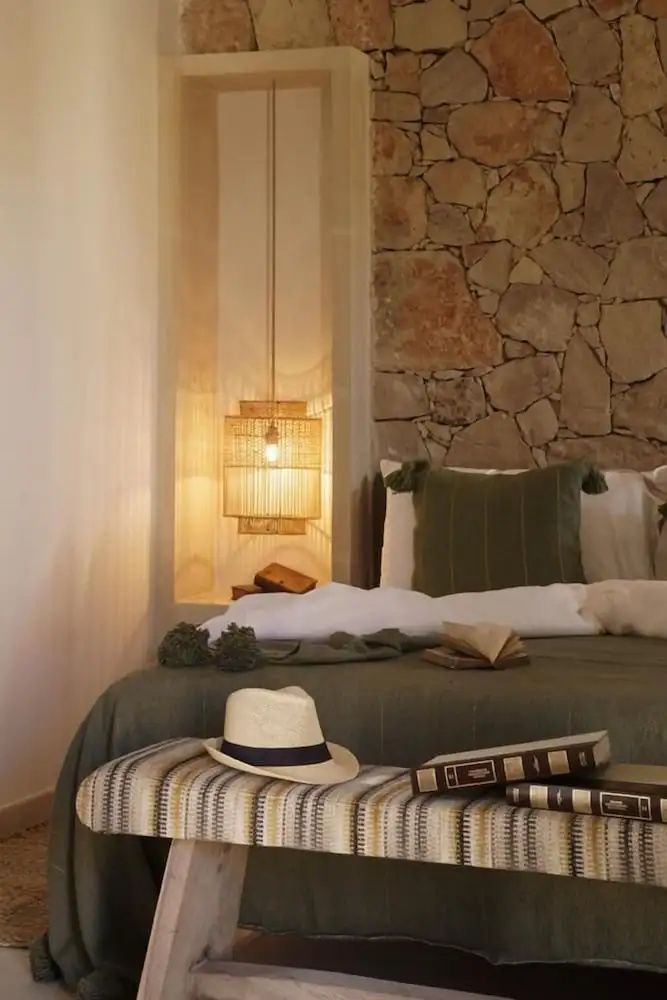Doppelbett mit grüner Tagesdecke, Natursteinwand, Strohlampe und einer Bank mit Strohhut.