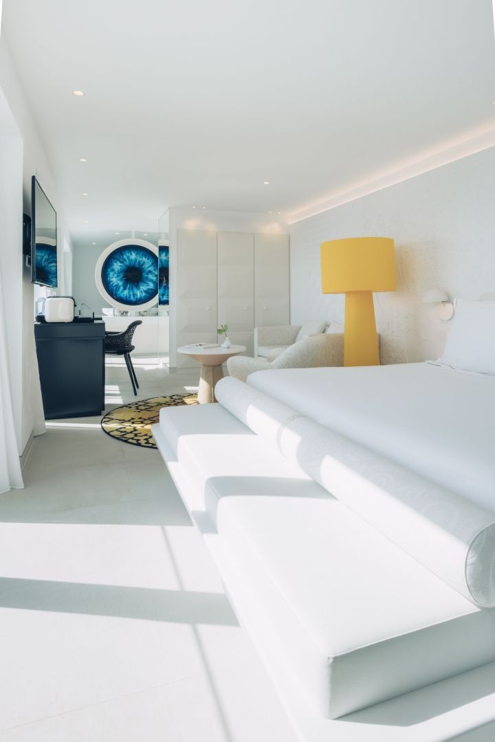 Weiß eingerichtetes Hotelzimmer mit weißem Bett, gelber Stehlampe und blauem Kunstwerk.