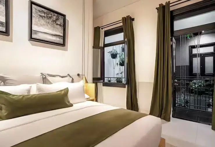 Hotelzimmer mit Doppelbett, grünen Vorhängen und einem geöffneten Fenster mit Blick auf Pflanzen.