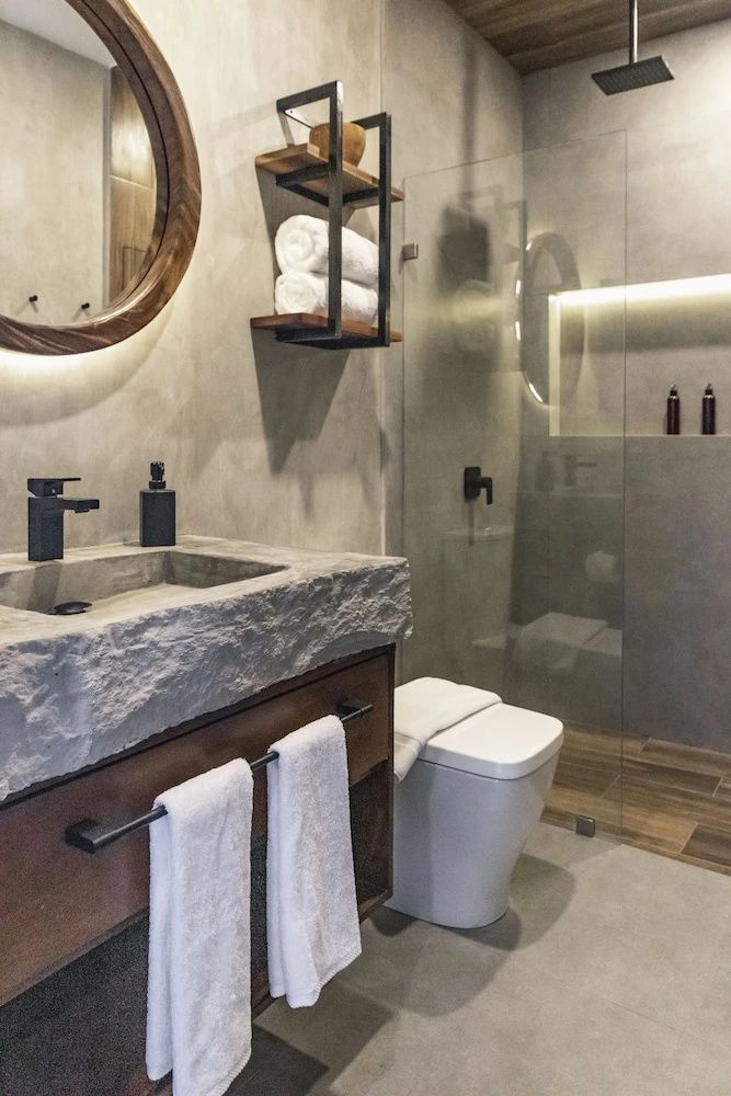 Badezimmer mit Naturstein-Waschtisch, rundem Spiegel, Toilette und einer Dusche mit Glaswand.