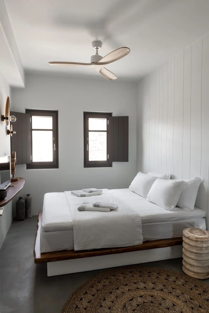 Helles Schlafzimmer mit Doppelbett, weißer Bettwäsche, zwei Fenstern mit Klappläden und Deckenventilator.