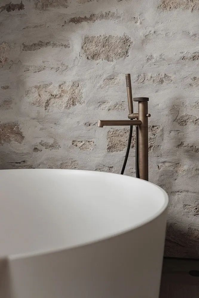 Freistehende weiße Badewanne mit bronzefarbener Armatur vor einer rustikalen Natursteinwand.