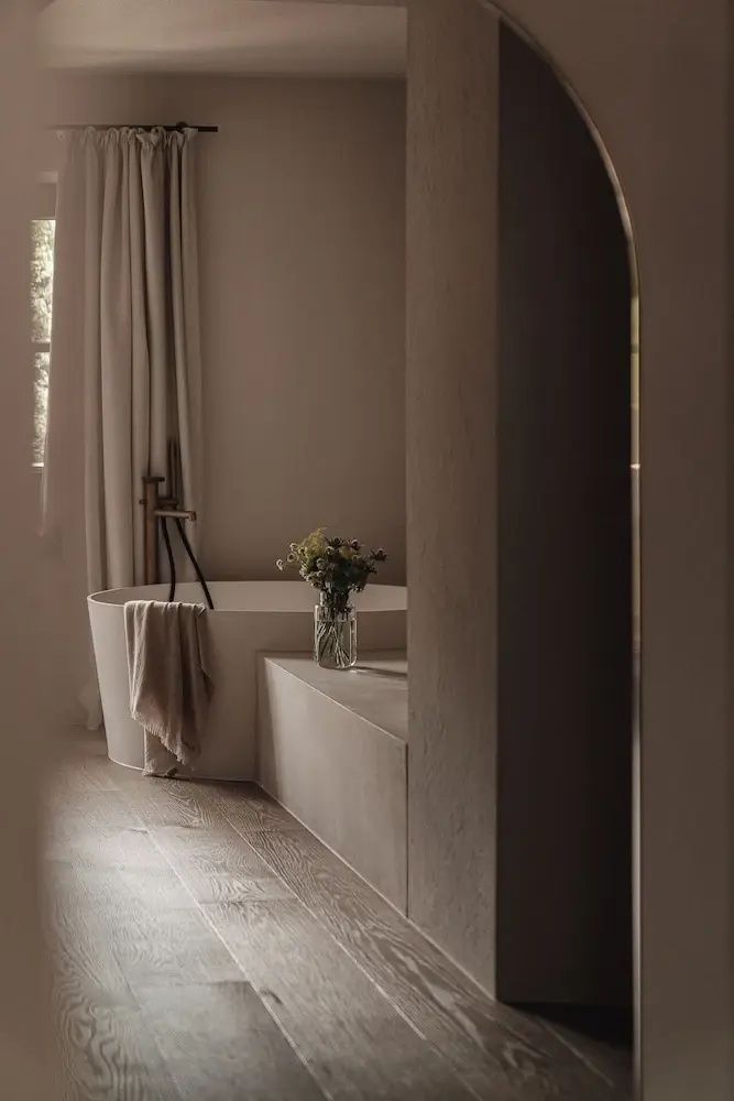 Freistehende runde Badewanne neben beigen Vorhängen und einer reduzierten Steinbank mit Blumendekoration.