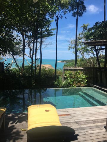 Ein Swimmingpool auf einer Holzterrasse umgeben von hohen Bäumen mit Meerblick.